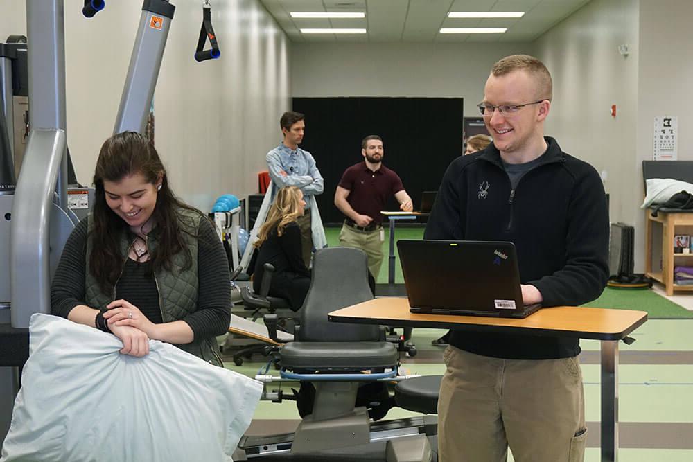 Male professor takes notes on laptop while observing female student in simulation lab