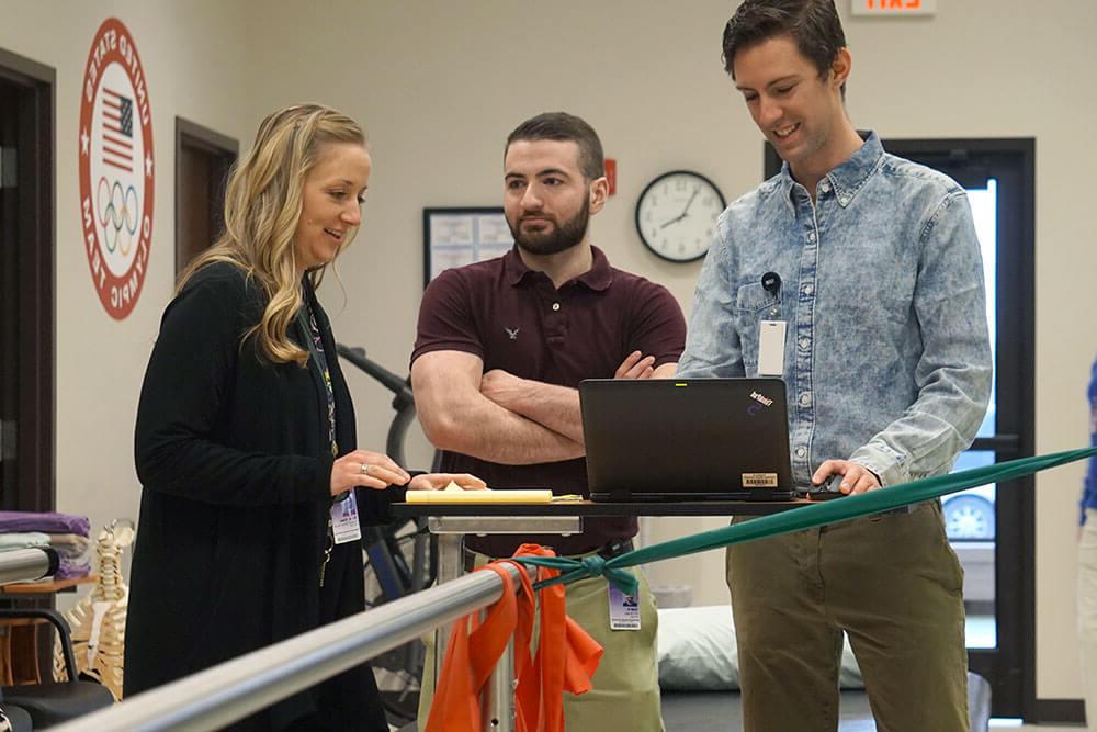 Two male students stand behind laptop while female professor gives them instructions