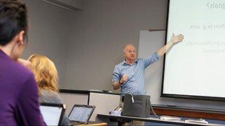 Professor points to screen as he lectures in front of students taking notes on laptops